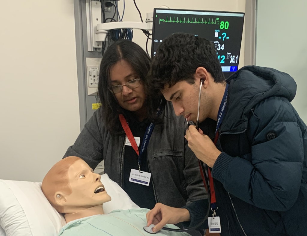 a young man using a stethoscope on a mannequin while a fellow student looks on